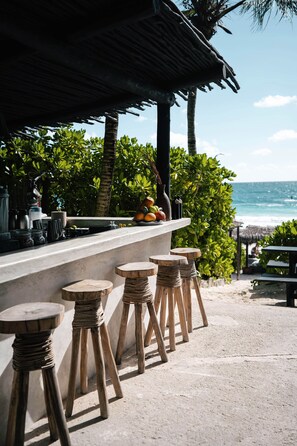 Un bar en la playa con taburetes de madera, una exhibición de frutas y vistas al océano.