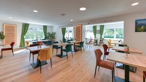 A modern dining area with wooden tables and chairs, large windows with green curtains, and a welcoming sign on the wall. - Pension Freiheit (Pfronten)