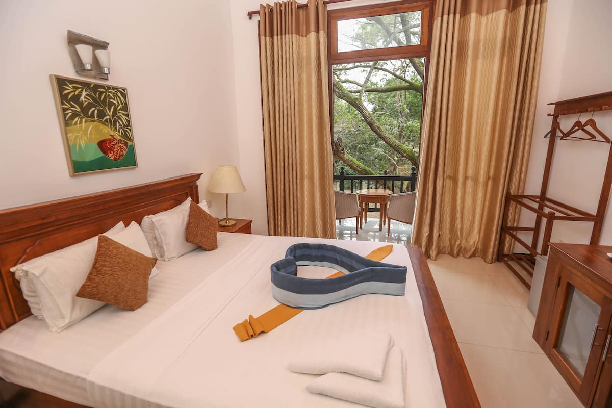 A neatly made bed with pillows, a blue neck pillow, and a folded towel. There is a wooden headboard, a bedside table with a lamp, and a window with curtains.