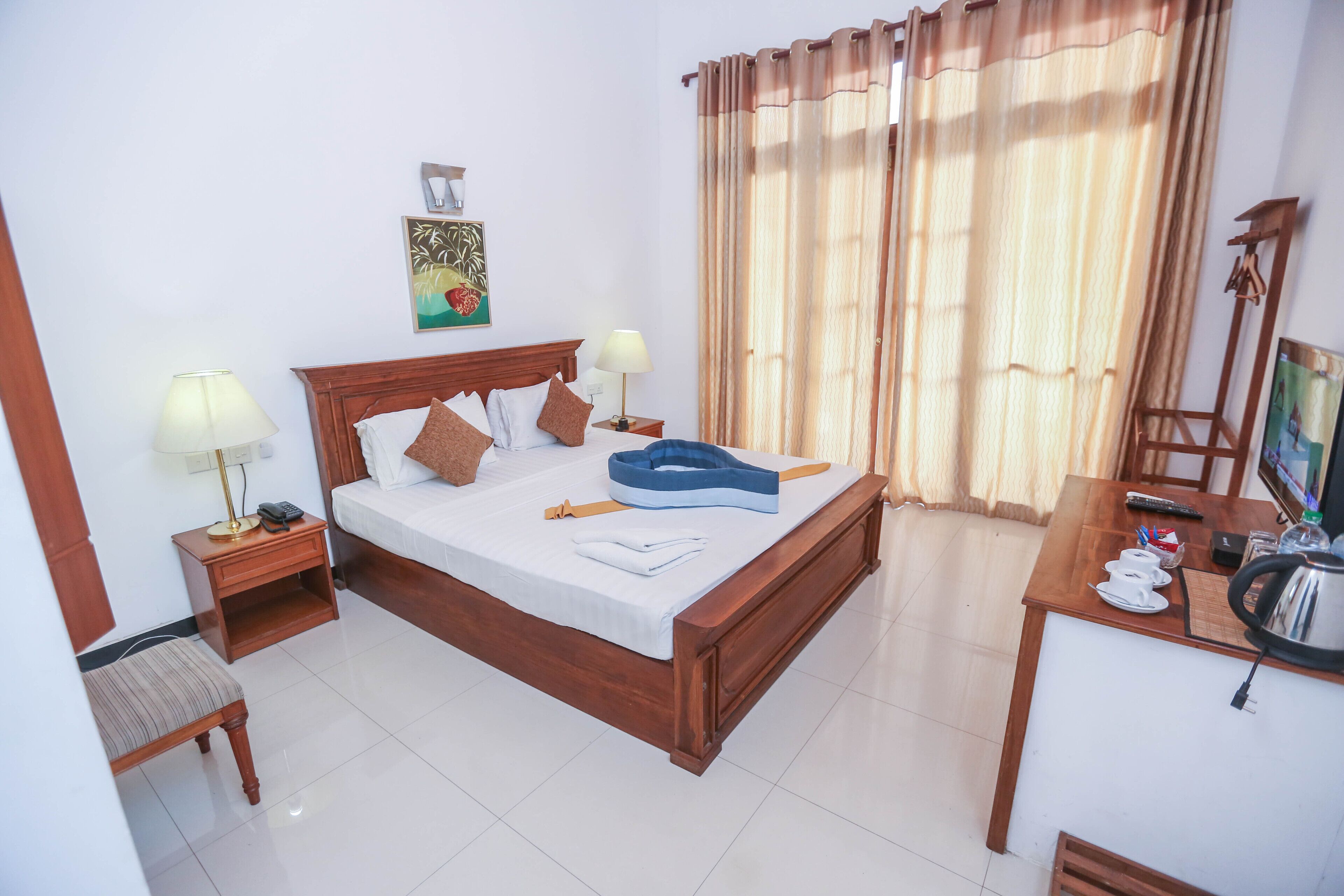 Habitación de hotel ordenada con una cama de madera, dos mesitas de noche con lámparas, un escritorio pequeño con un hervidor y una ventana con cortinas.