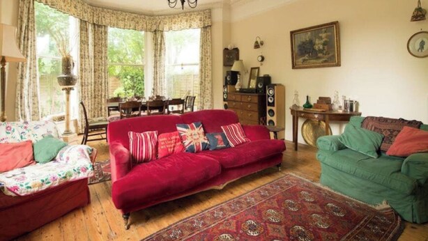 Una sala de estar acogedora con un sofá rojo, un sofá verde y un sofá estampado con motivos florales. Se aprecia un ventanal con cortinas, una araña y un cuadro en la pared.