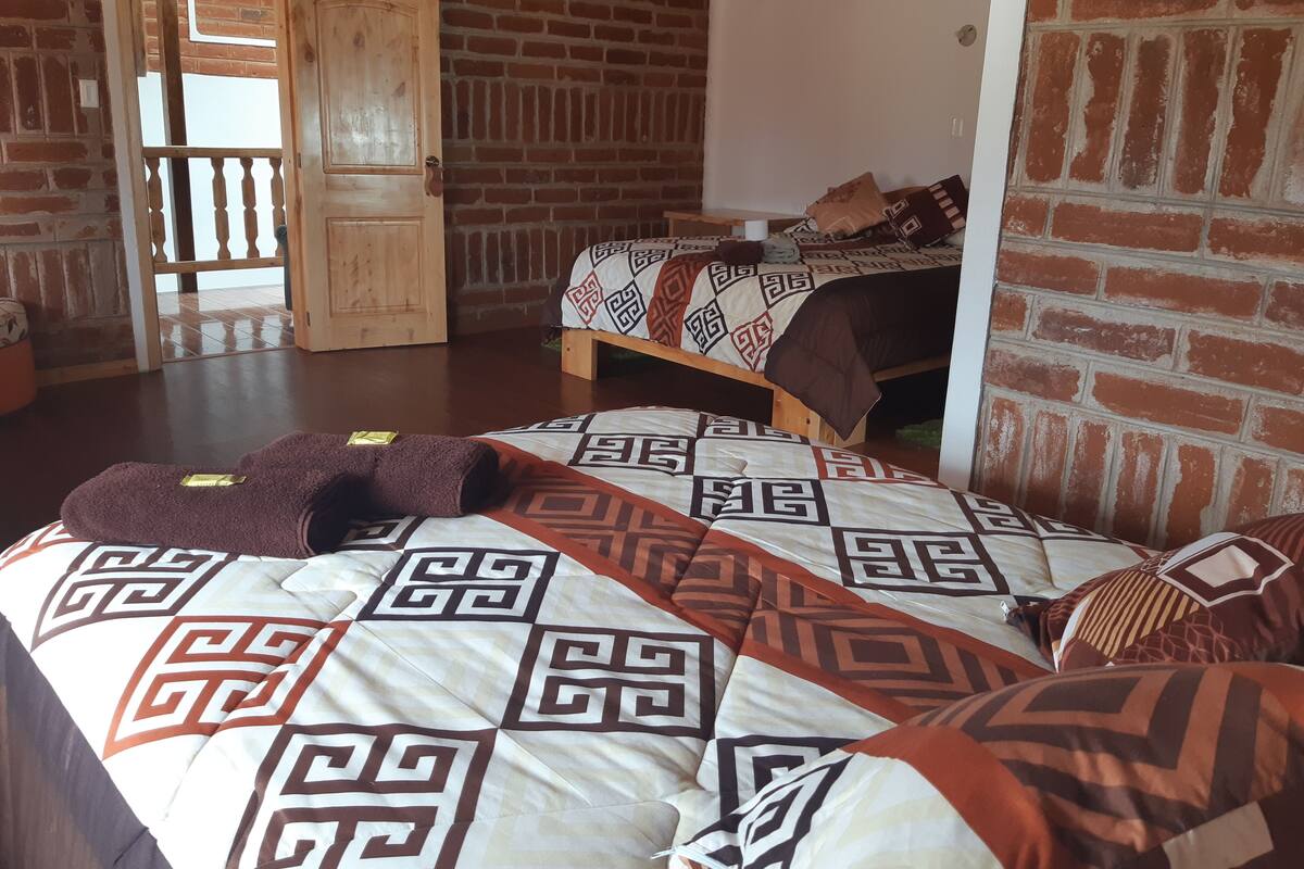 A room with a brick wall, a wooden door, a bed with patterned bedding, and a small table with a cup.