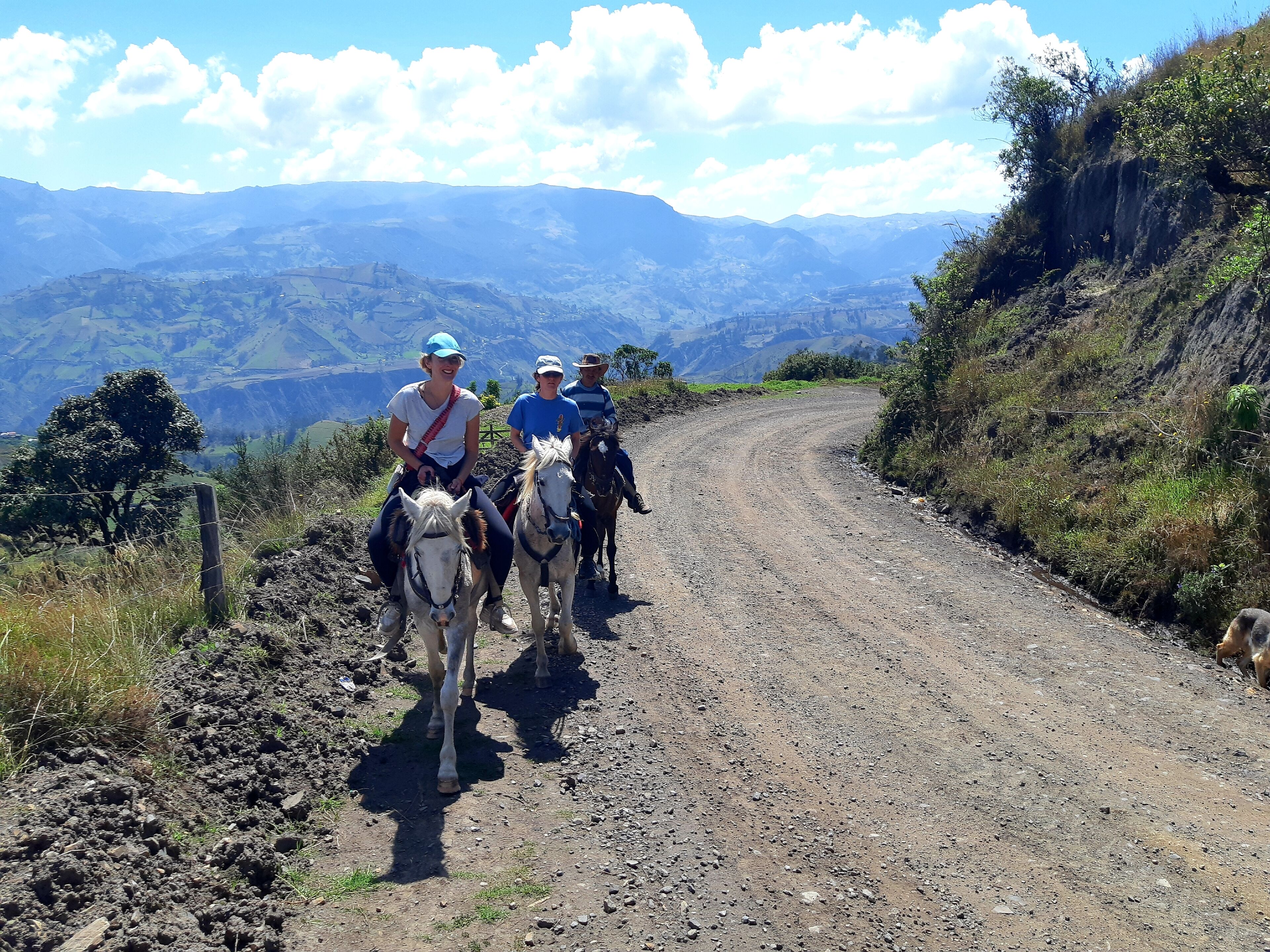 Three people riding horses on a dirt road with mountainous terrain and sparse vegetation on either side.