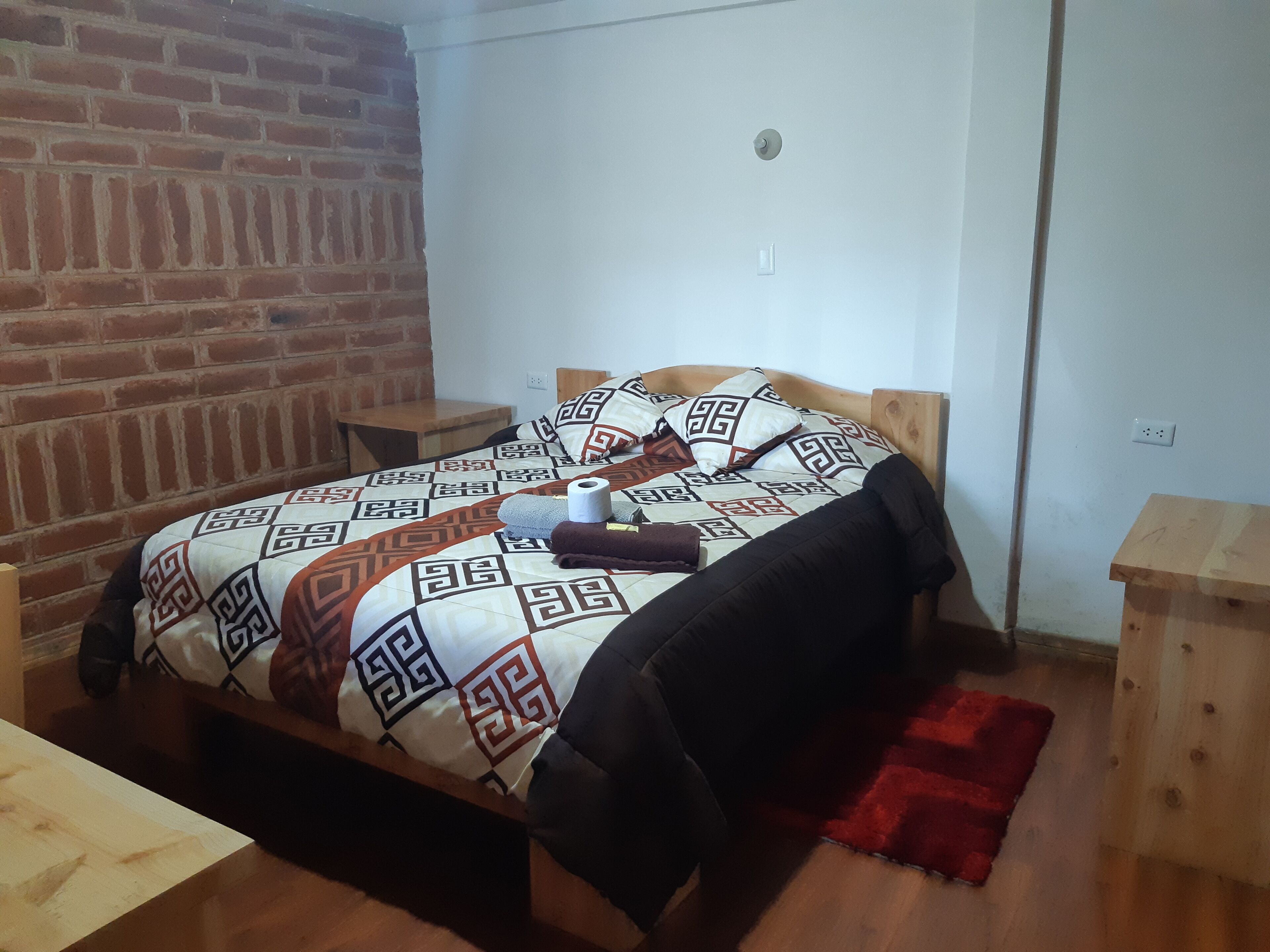 A single bed with a patterned bedspread, a wooden headboard, and a red rug on the floor. There are two bedside tables, one with a lamp and the other with a power outlet.