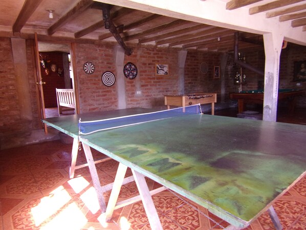 Una mesa de ping pong en una habitación con pared de ladrillo y vigas de madera. Hay dardos en la pared y una mesa de fútbol de mesa al fondo.