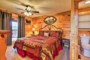Une chambre dans une cabane en bois, avec un lit, un ventilateur de plafond, une table de chevet avec une lampe, une tête de lit en bois, un miroir et une fenêtre avec des rideaux.