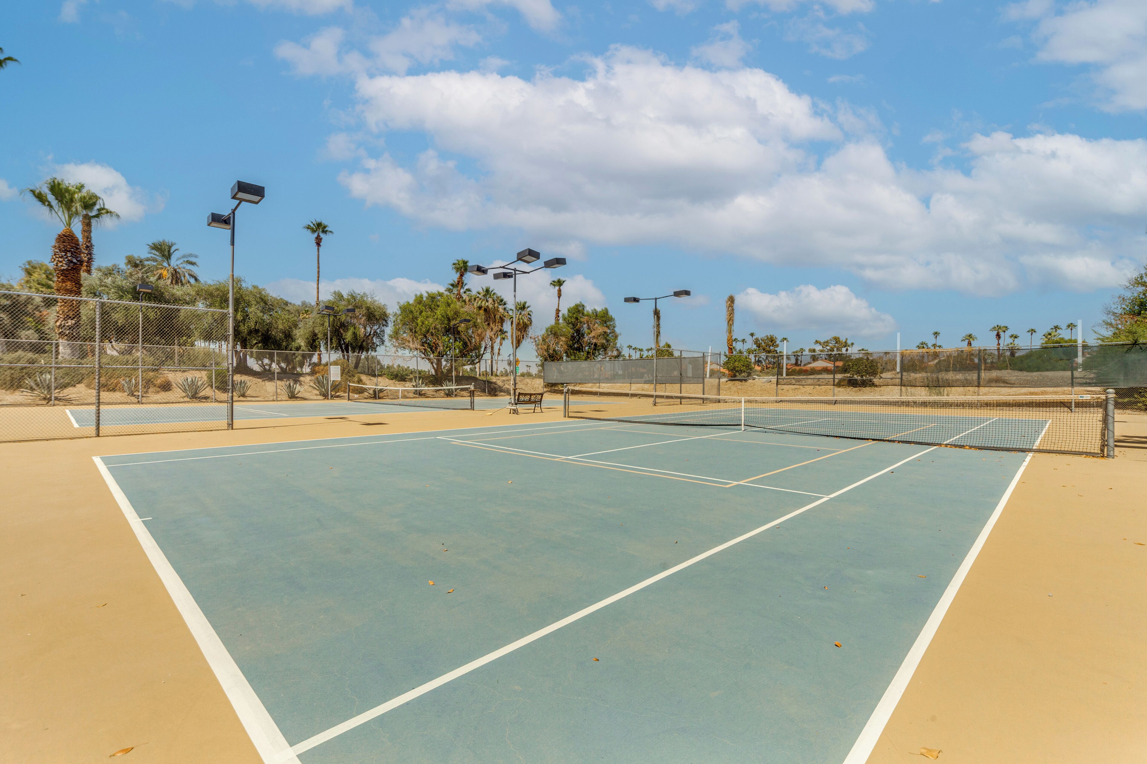 Un campo da tennis all'aperto con una rete, circondato da una recinzione e palme sullo sfondo.