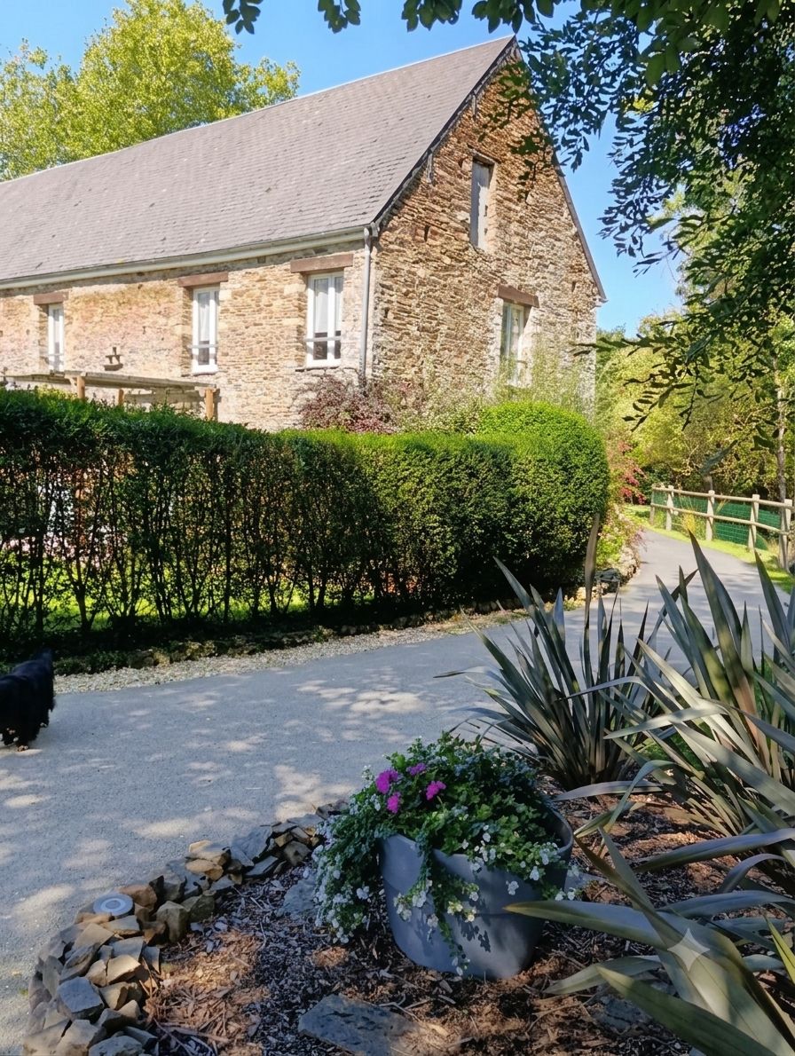 Een gebouw van steen met een schuin dak, een verharde oprit en een verzorgde tuin met diverse planten en een haag.