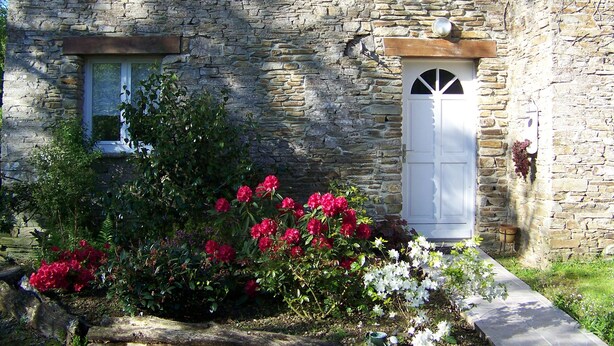 A stone building with a white door and window, surrounded by flowering plants.