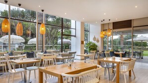 Ein modernes Restaurant mit Holztischen und weißen Stühlen, großen Fenstern mit Sonnenschirmen und hängenden Pendelleuchten.