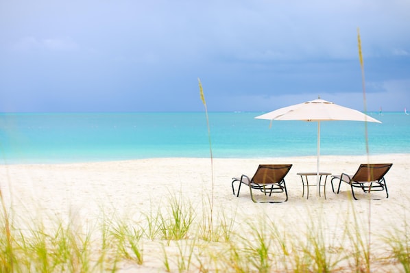 A beach with two chairs and a table under an umbrella, facing the ocean.