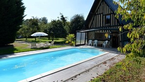 En swimmingpool med træterrasse, omgivet af en have og en bygning med skråt tag.