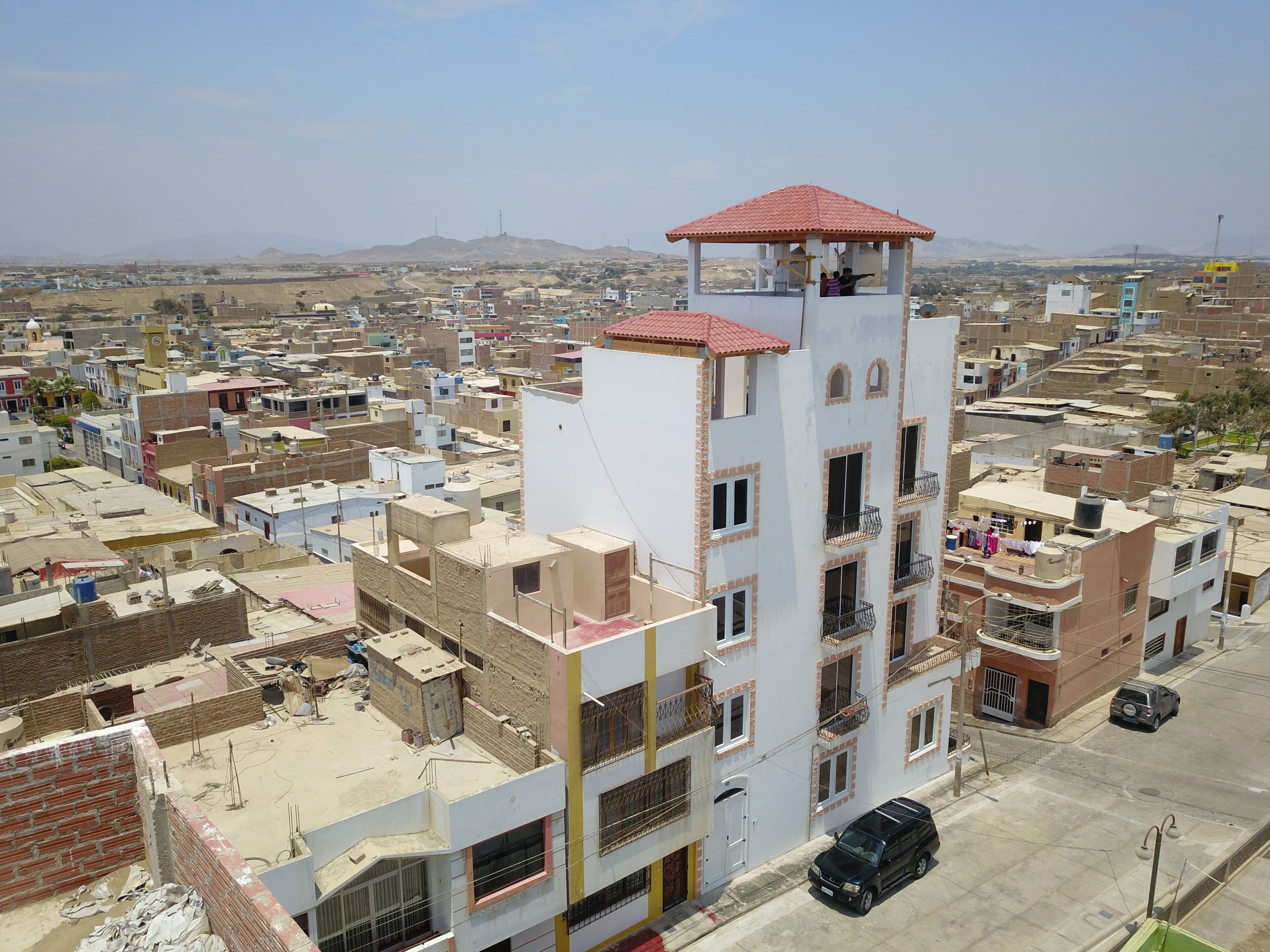Un edificio de varios pisos con techo de tejas rojas, rodeado de una zona urbana densamente poblada con diversos edificios y una cadena montañosa en la distancia.
