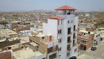 Un edificio de varios pisos con techo de tejas rojas, rodeado de una zona urbana densamente poblada con diversos edificios y una cadena montañosa en la distancia.