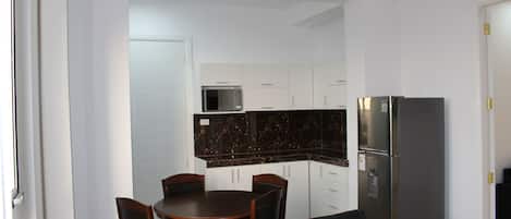 Una cocina compacta con armarios blancos, un protector contra salpicaduras de mármol oscuro, un refrigerador y una zona de comedor con mesa y sillas.