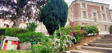 Un jardín con una casita de juegos pequeña, un árbol alto y un edificio con franjas rojas y blancas.