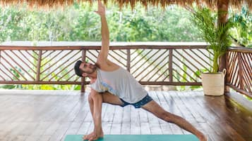 Una persona realizando una postura de yoga en una terraza de madera con techo de paja, rodeada de follaje.