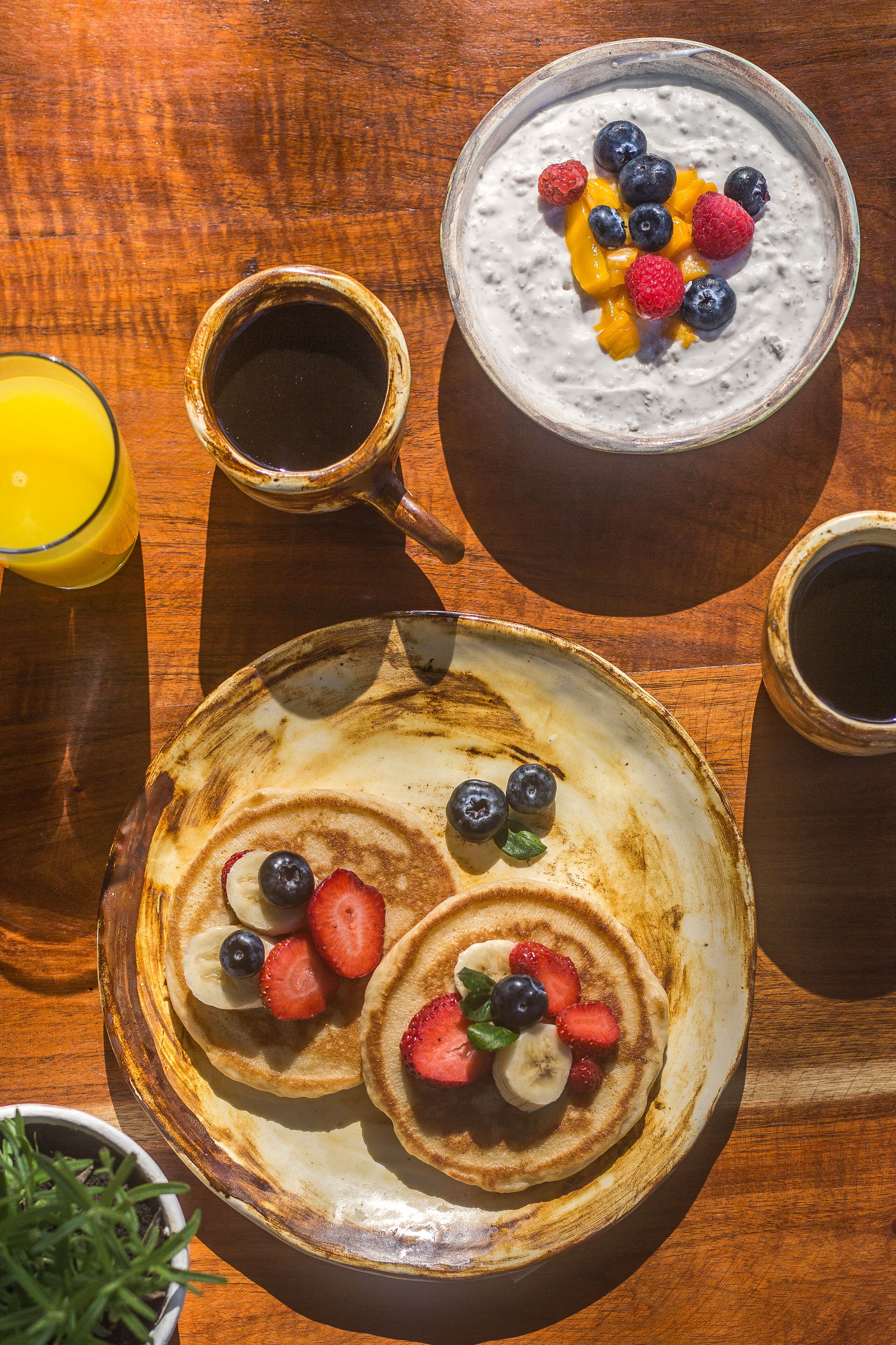 Un desayuno con tortitas, yogur y café sobre una mesa de madera.