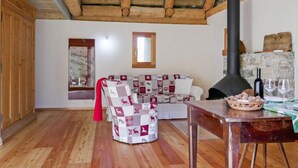 A cozy living room with a wooden ceiling, a stone fireplace, a sofa with patterned cushions, and a dining table with a bowl of snacks. - Rustico Andrea by Interhome (Castro)