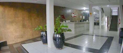 A hotel lobby with a reception desk, potted plants, and a tiled floor.