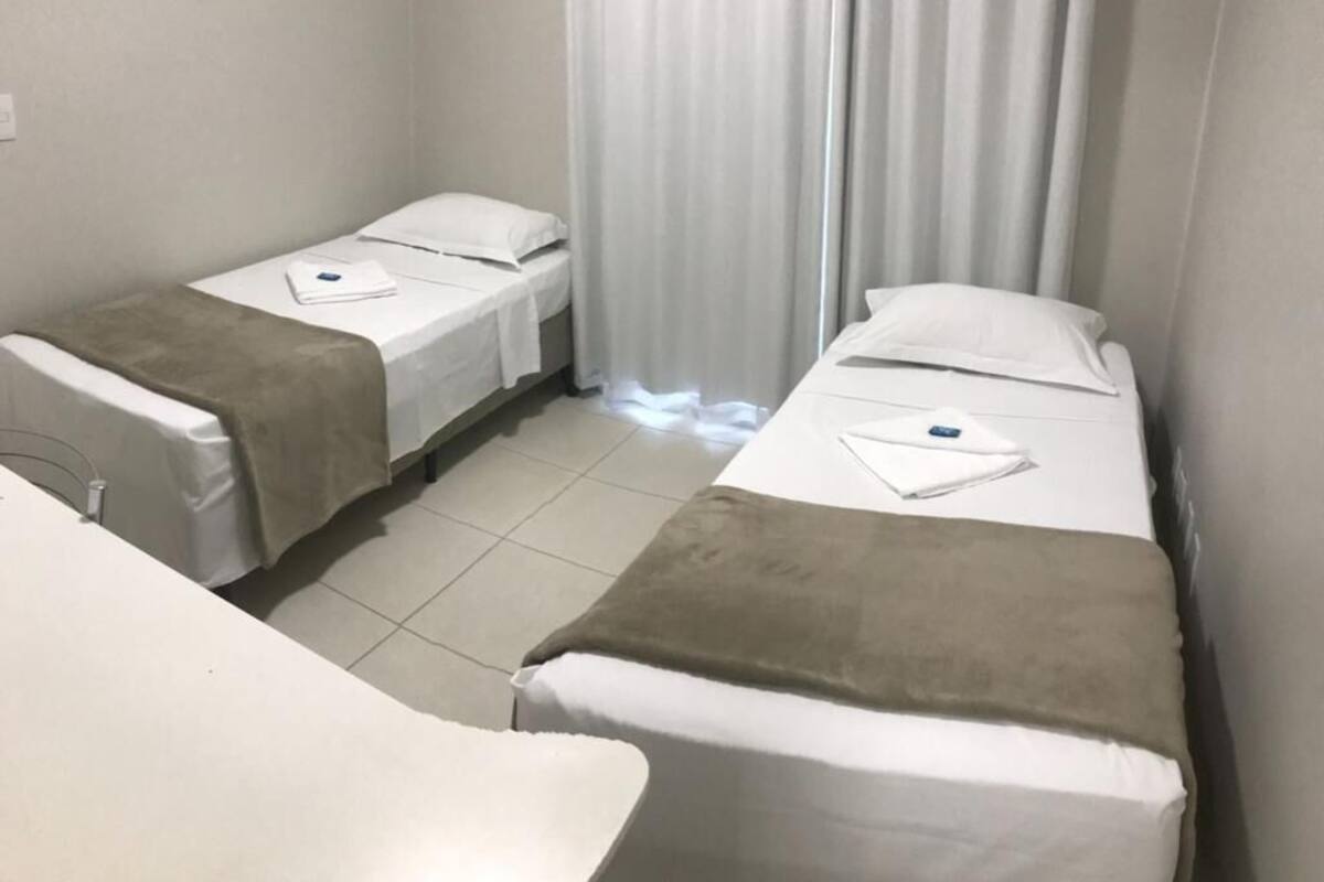Habitación de hotel con dos camas individuales, cada una con un colchón blanco y una manta marrón. Hay una ventana con cortinas blancas, y el piso es de cerámica.