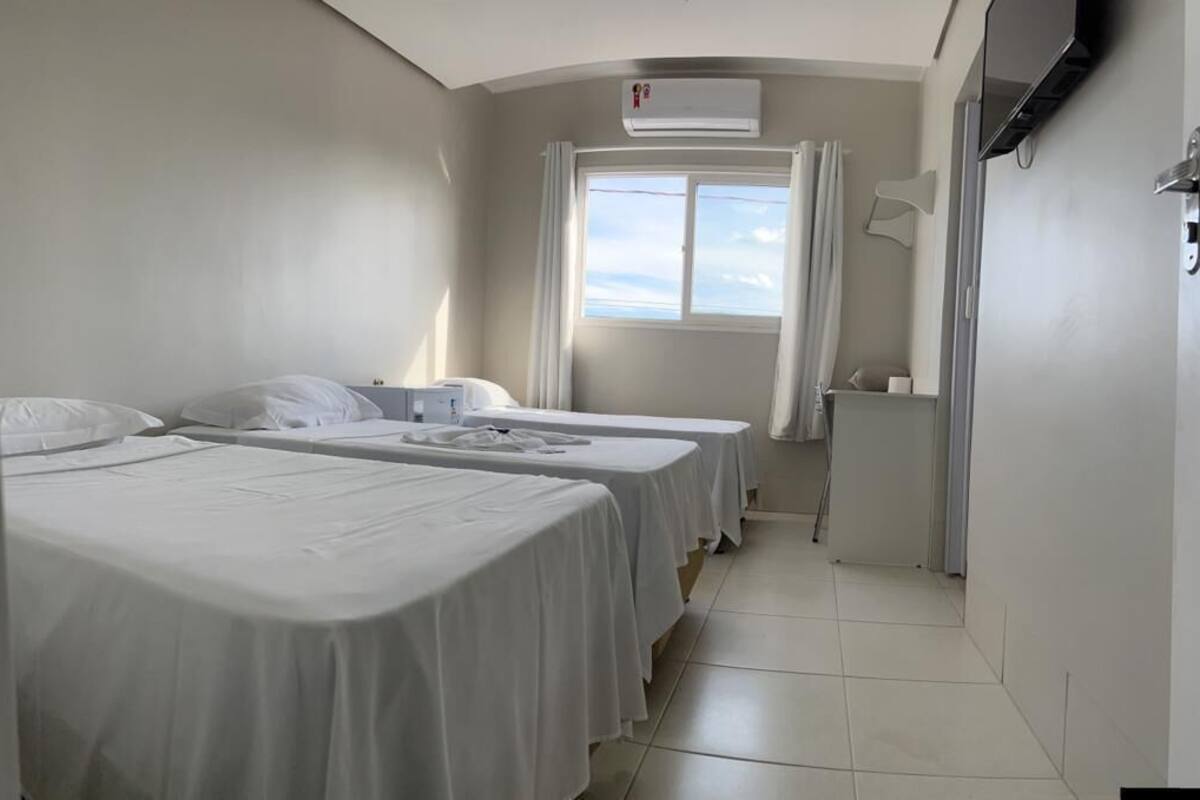 Habitación de hotel con dos camas individuales, una ventana con cortinas, un equipo de aire acondicionado y un televisor fijado a la pared.