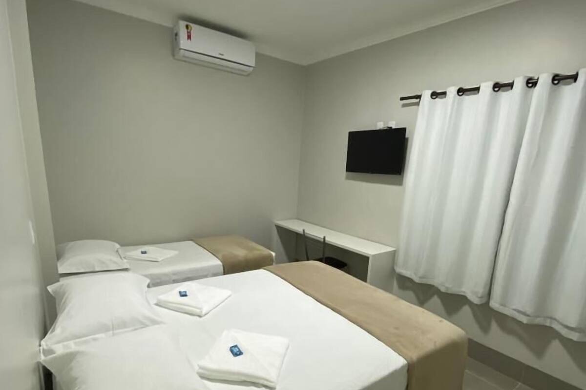 Una habitación de hotel con cama, mesita de noche, televisor y ventana con cortinas.