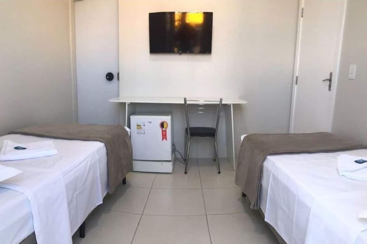 Una habitación con dos camas individuales, un escritorio pequeño con silla, un refrigerador y un televisor en la pared.