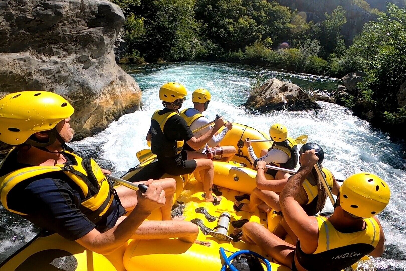 A group of people in a yellow raft navigating a river surrounded by rocky terrain and greenery.