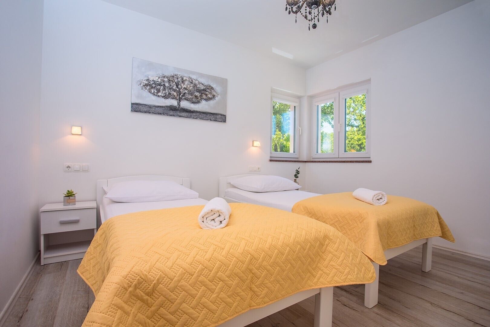 A hotel room with two beds, each with a yellow quilted bedspread and a rolled towel. There is a painting of a tree on the wall, a bedside table with a plant, and a window with a view of greenery.