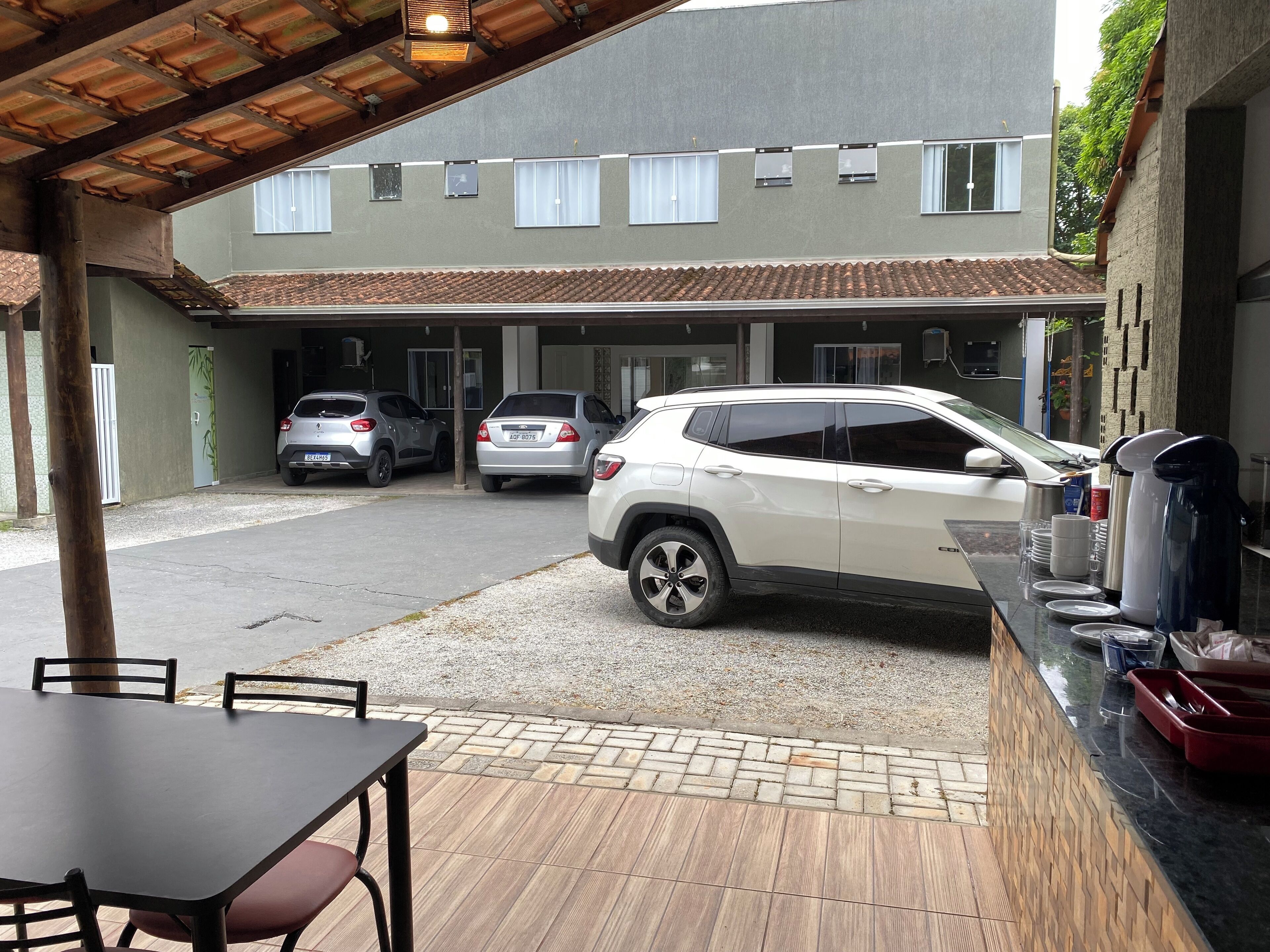 Un espacio de estar techado al aire libre con mesa y sillas, un estacionamiento con tres coches, y un edificio con tejado de tejas y ventanas.
