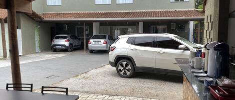 Un espacio de estar techado al aire libre con mesa y sillas, un estacionamiento con tres coches, y un edificio con tejado de tejas y ventanas.