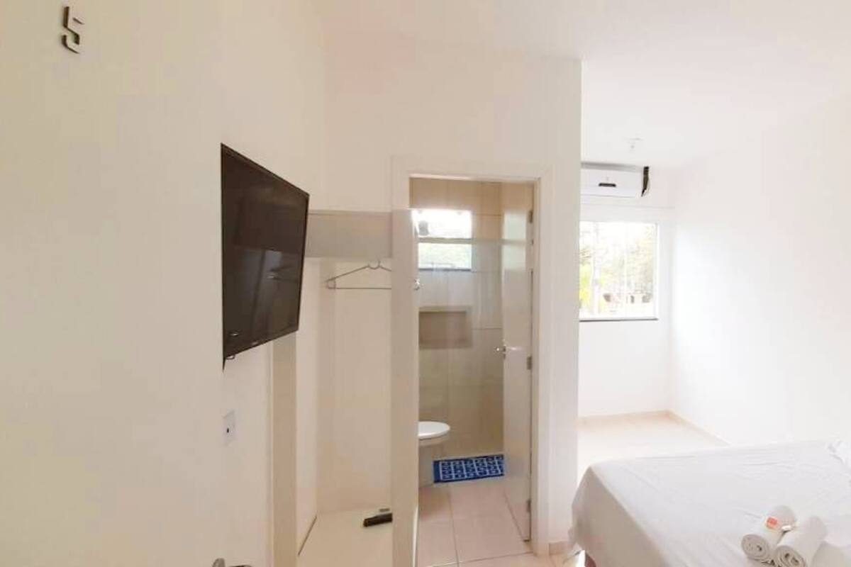 A hotel room with a bed, a TV mounted on the wall, a bathroom with a toilet and a shower, and a window providing natural light.