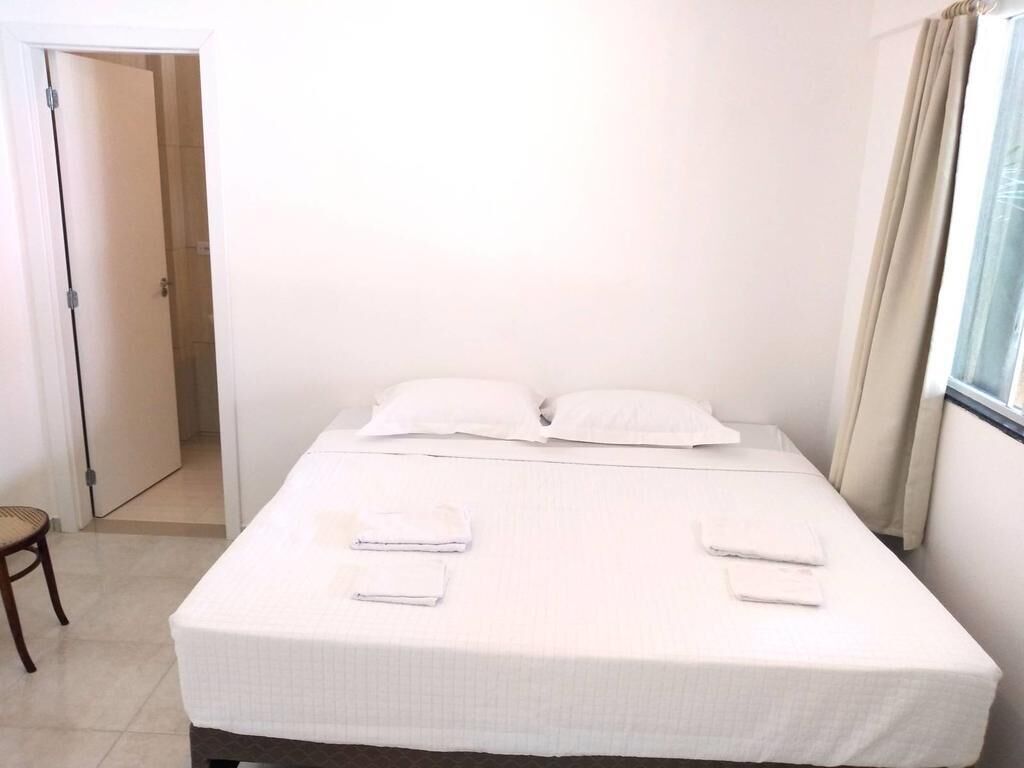 Una cama bien hecha con sábanas y almohadas blancas, una silla de madera y una puerta que da a otra habitación.