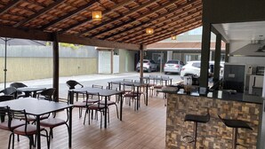 An outdoor dining area with wooden tables and chairs, a tiled floor, and a kitchen counter with stools. - Pousada Pontal do Sul (Pontal do Parana)