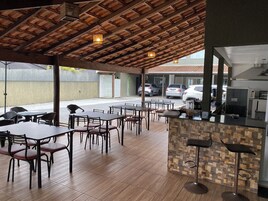 Un área de comedor al aire libre con mesas y sillas de madera, piso de baldosas y una barra de cocina con taburetes.