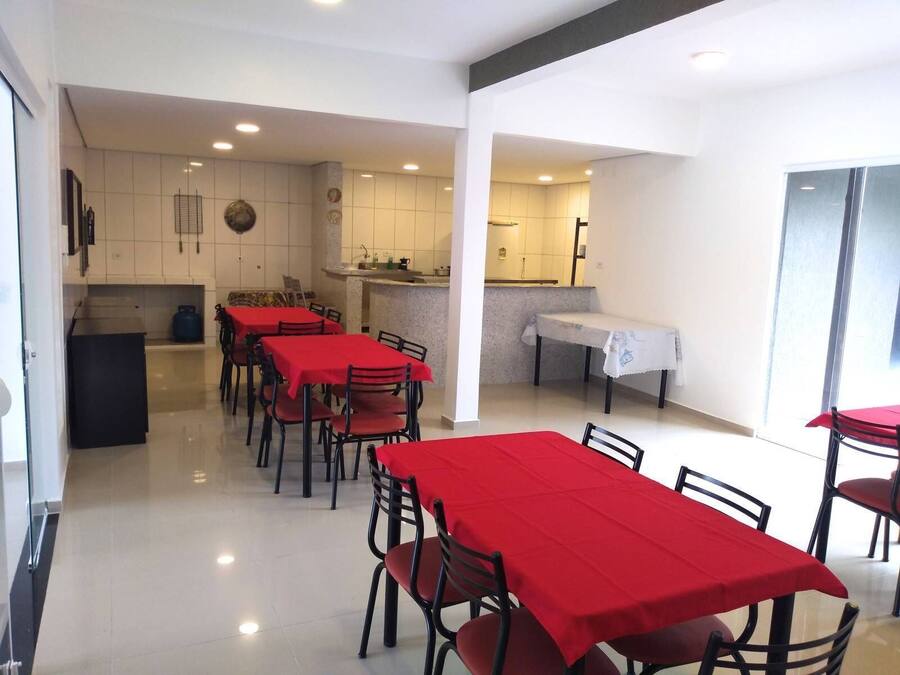 A dining area with red tablecloths and chairs, a kitchen in the background, and a large window providing natural light.