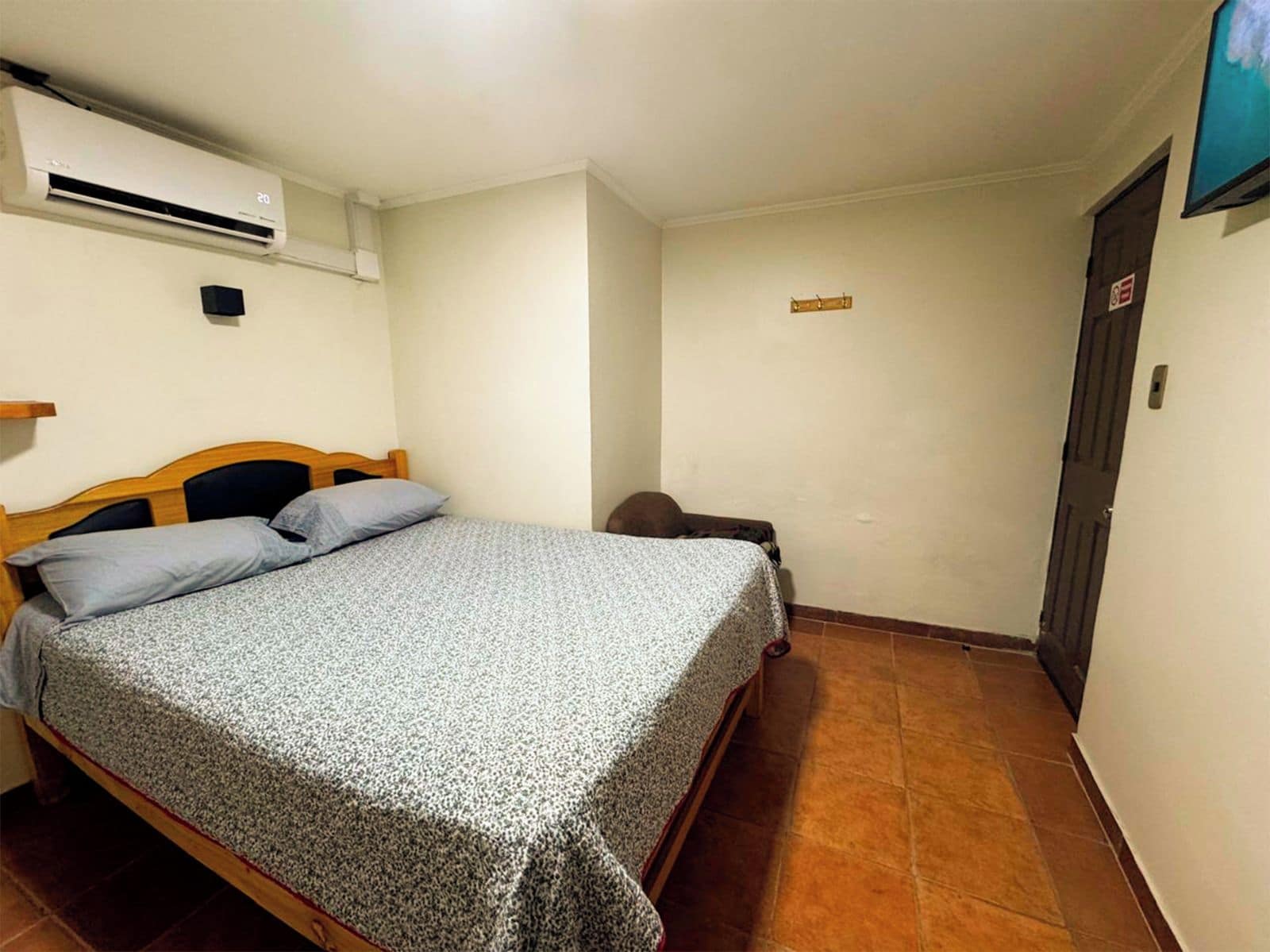 A single bed with a patterned bedspread, a wall-mounted air conditioner, a small bedside table, and a door with a sign.
