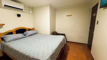 A single bed with a patterned bedspread, a wall-mounted air conditioner, a small bedside table, and a door with a sign.