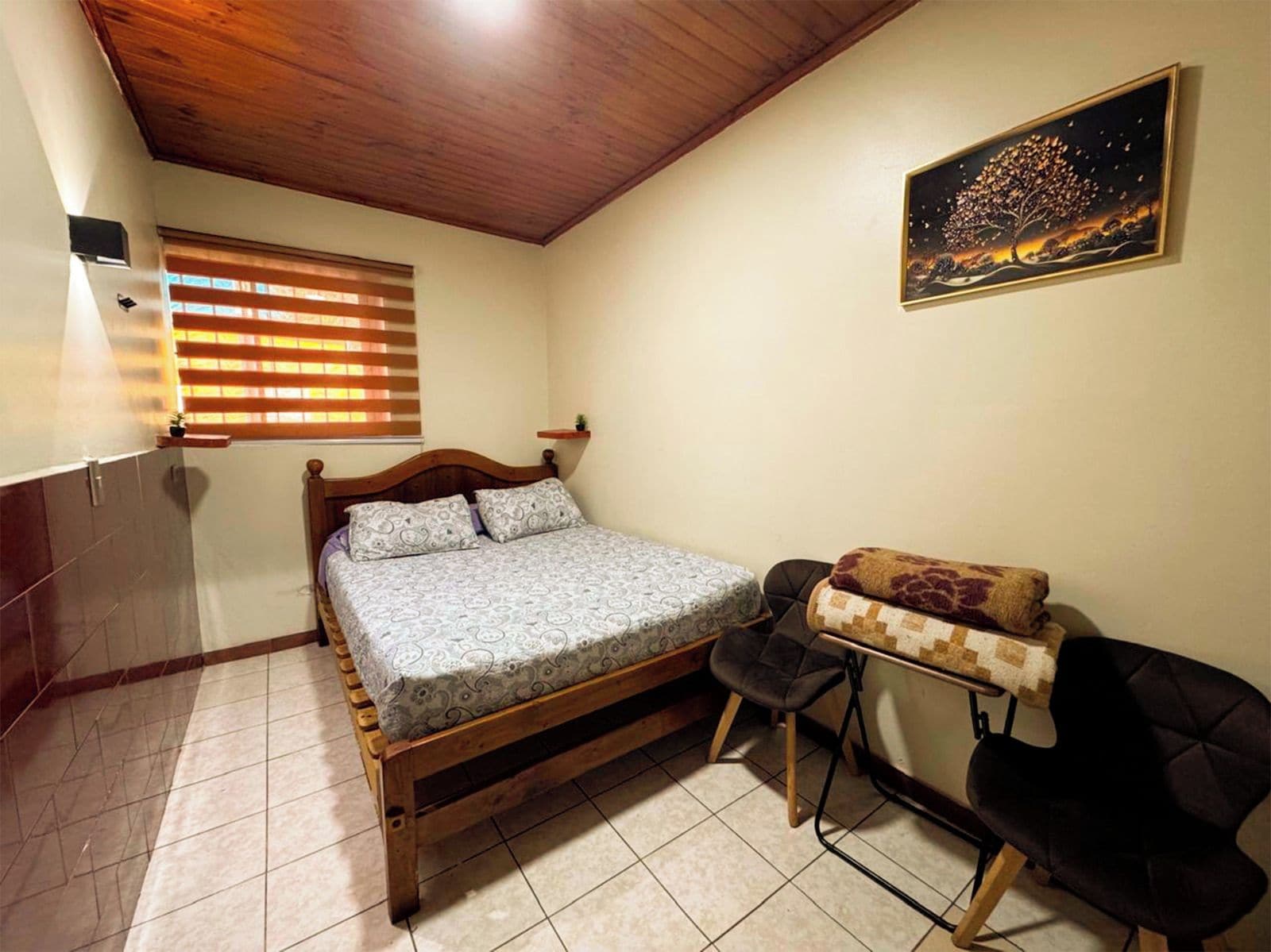 Un dormitorio con una cama de madera, un escritorio pequeño, una silla, una ventana con persianas y un cuadro en la pared.