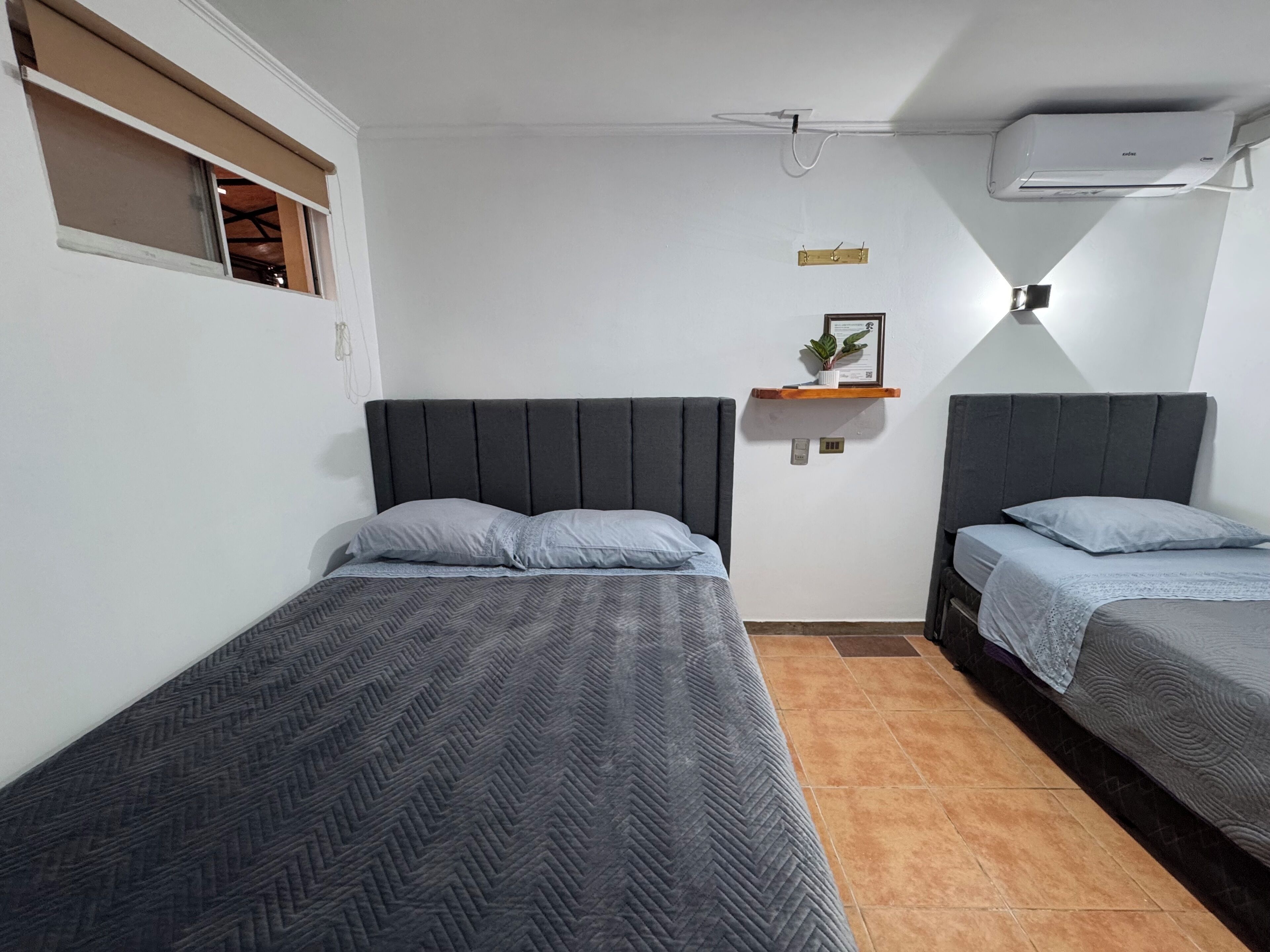 A room with two single beds, a small shelf with a plant, a wall-mounted air conditioner, and a window with a wooden blind.