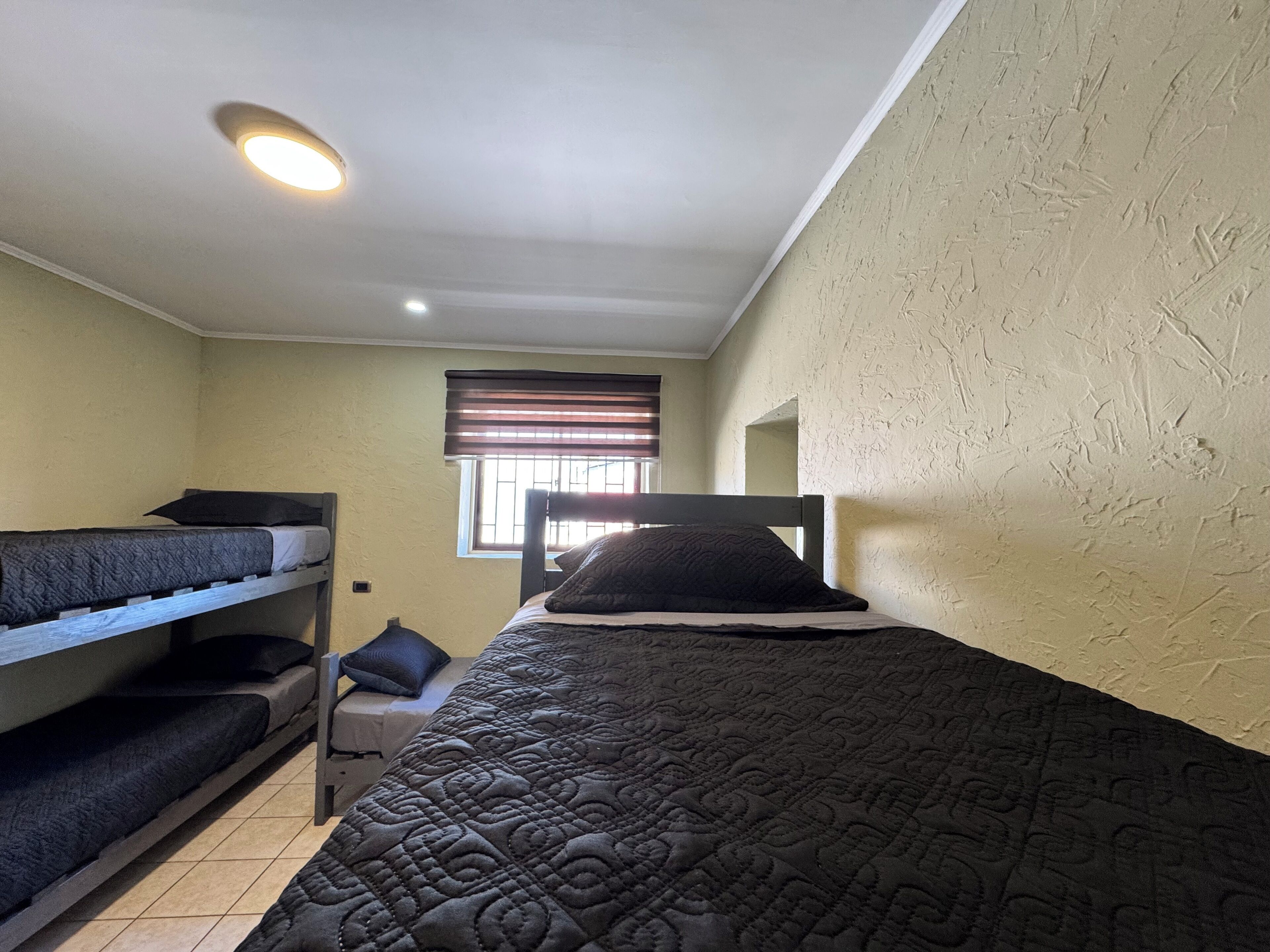 A dormitory room with bunk beds, a window with blinds, and a ceiling light.
