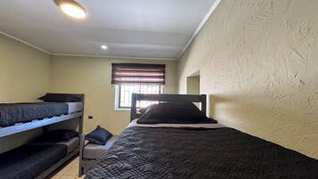 A dormitory room with bunk beds, a window with blinds, and a ceiling light.