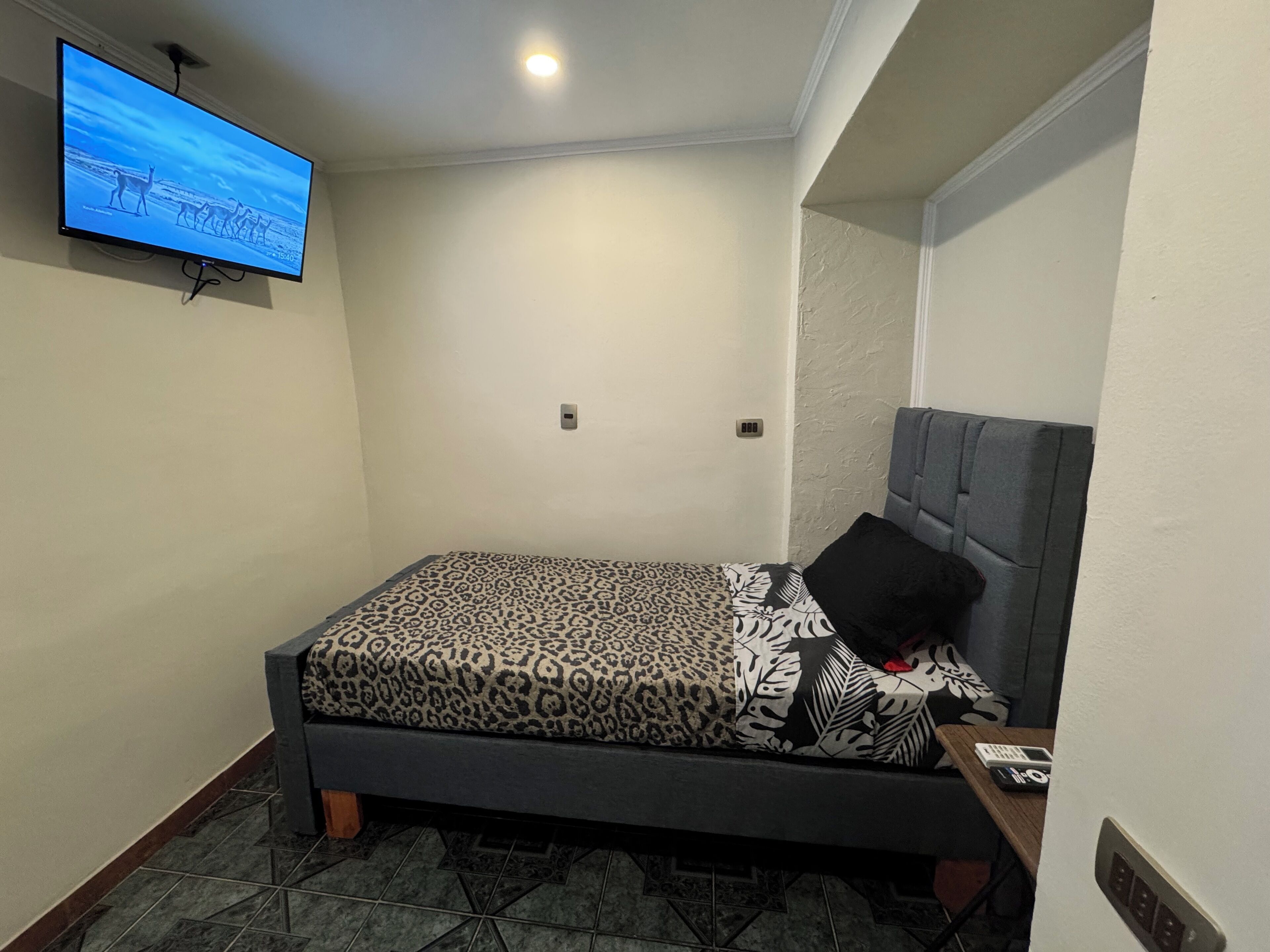 A small, neatly arranged room with a bed, a television mounted on the wall, and a door with a handle.