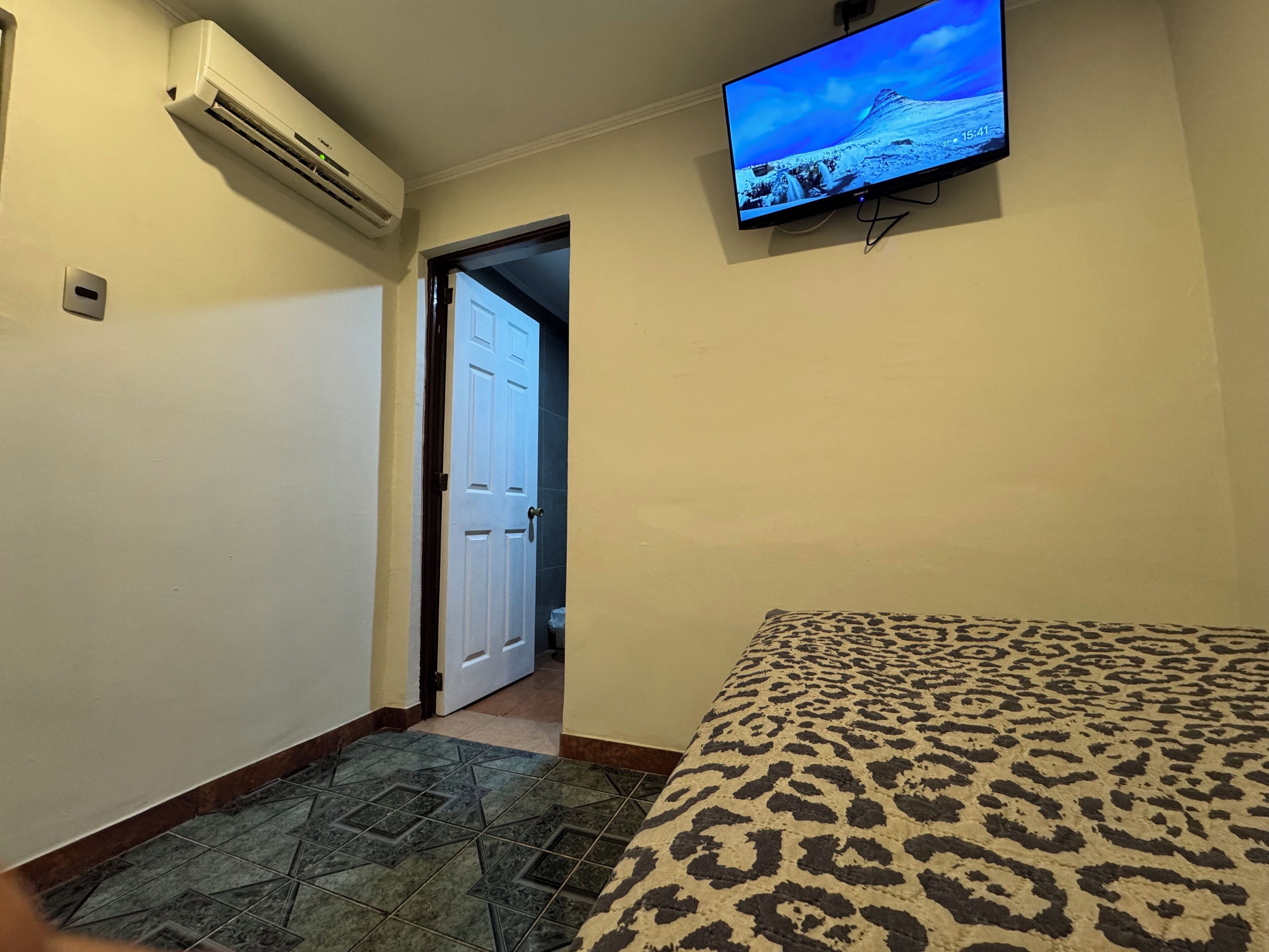 Una habitación de hotel con una cama, un televisor montado en la pared y un equipo de aire acondicionado.