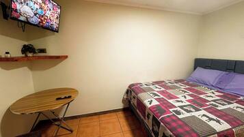 Un dormitorio con cama, televisor montado en la pared, una mesita y una estantería con una planta.