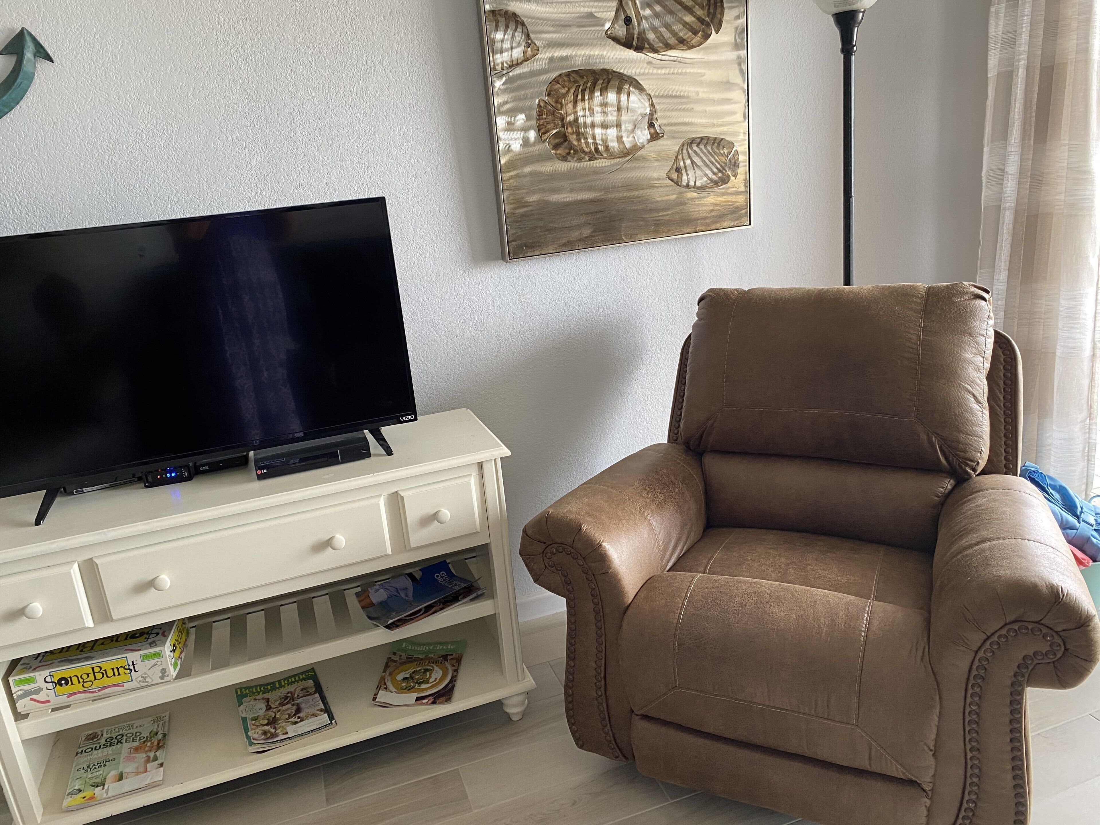 A living room with a flat-screen TV on a white cabinet, a brown leather armchair, and a wall art piece with shells.