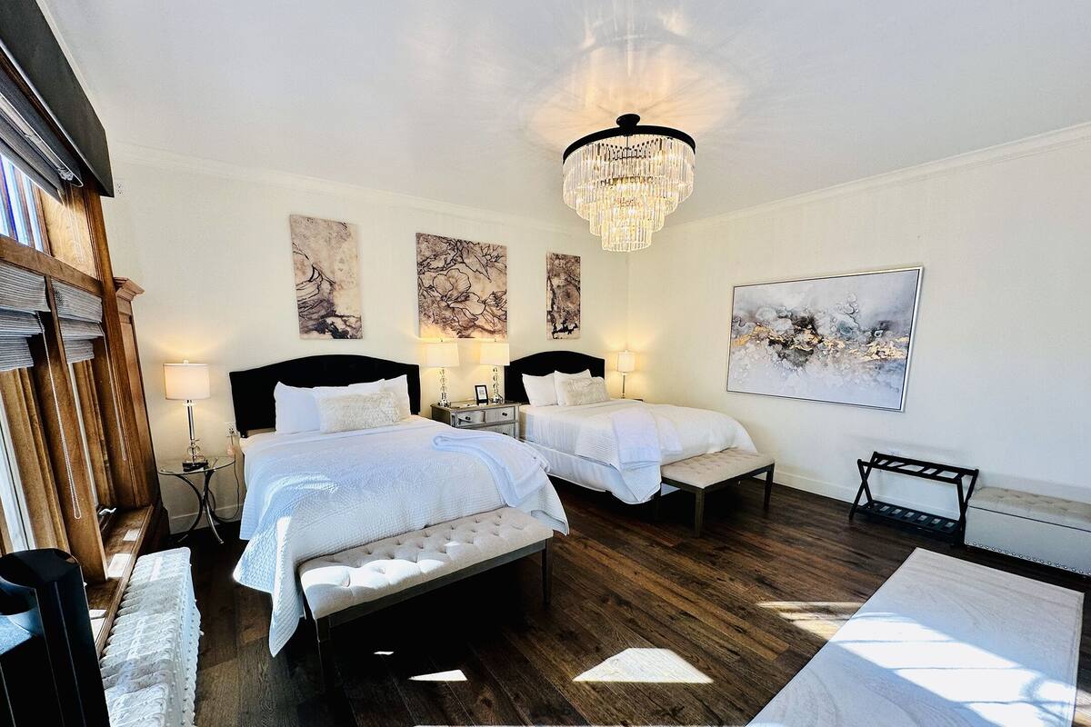 A hotel room with two beds, a chandelier, artwork on the walls, and a bench.