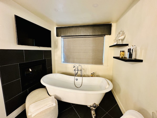A bathroom with a freestanding bathtub, a toilet, a towel, a window with a blind, and a shelf with toiletries. - Bespoke Inn Flagstaff (Flagstaff)