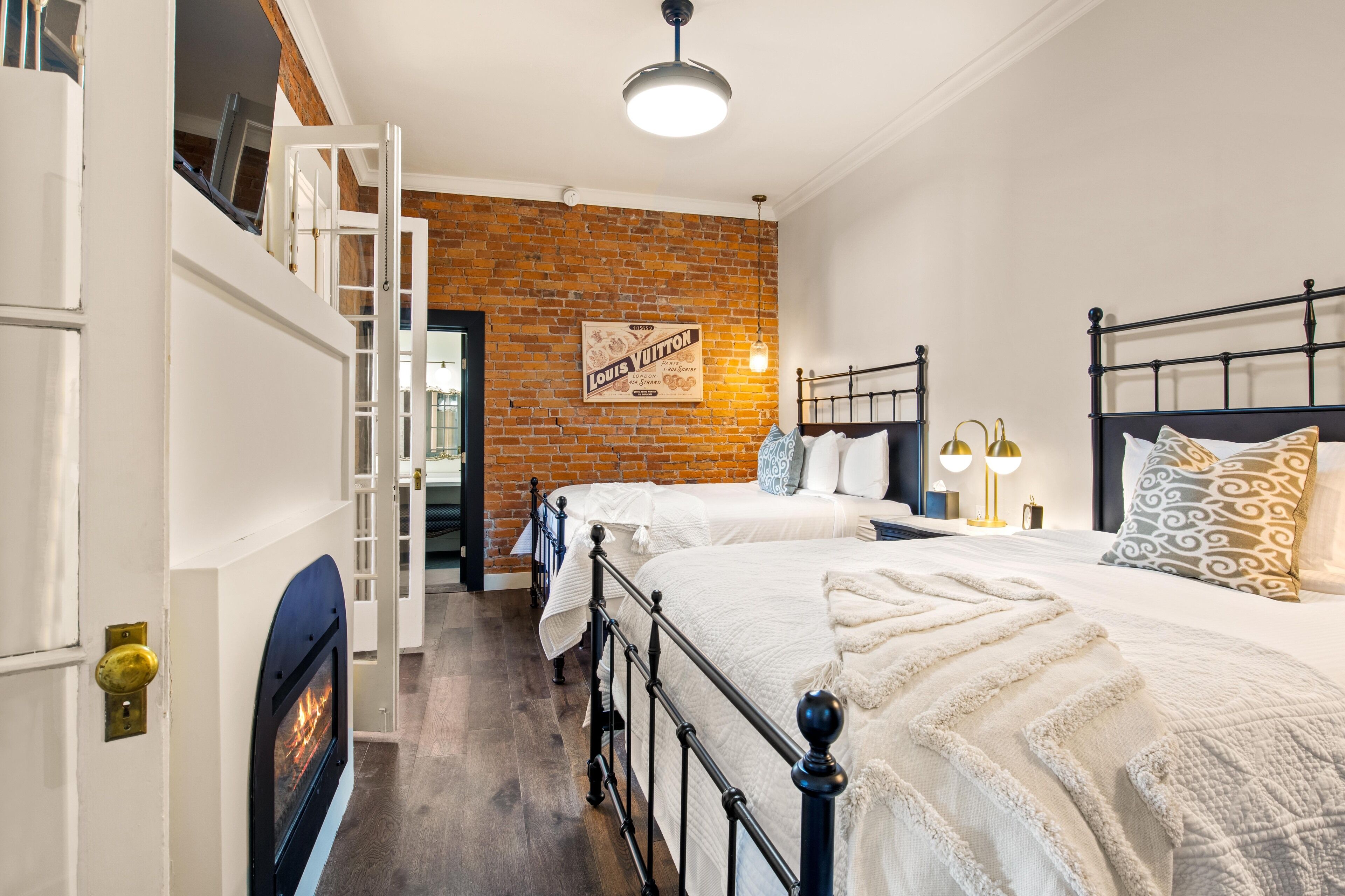 A bedroom with two beds, a brick wall, a Louis Vuitton poster, and a fireplace.